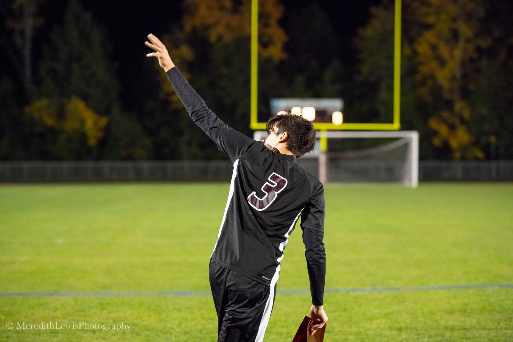 A soccer player in a number 3 jersey raising an arm on the field under the lights at dusk.