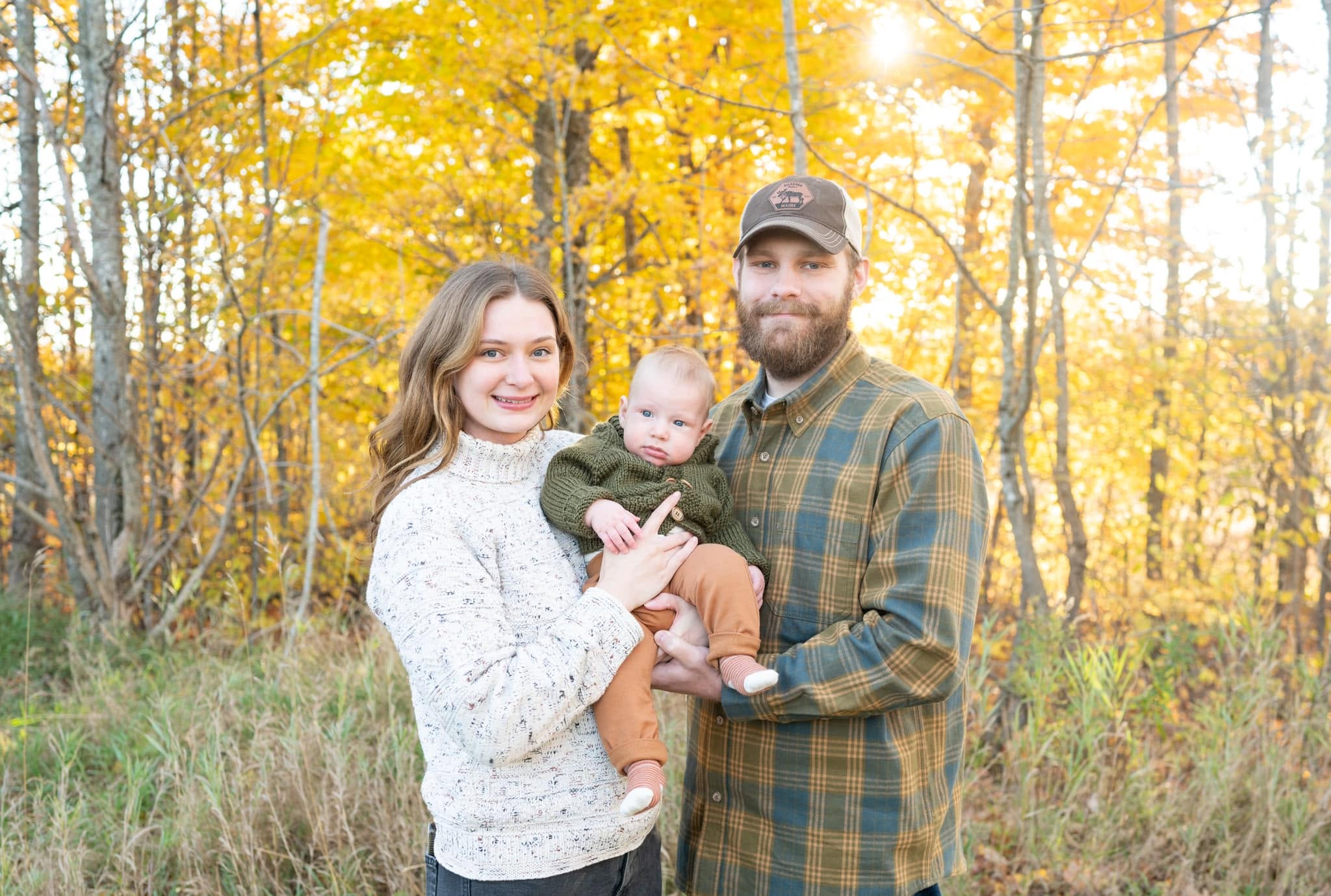 A mother and father holding their baby in front of golden autumn birch trees backlit by the sun.